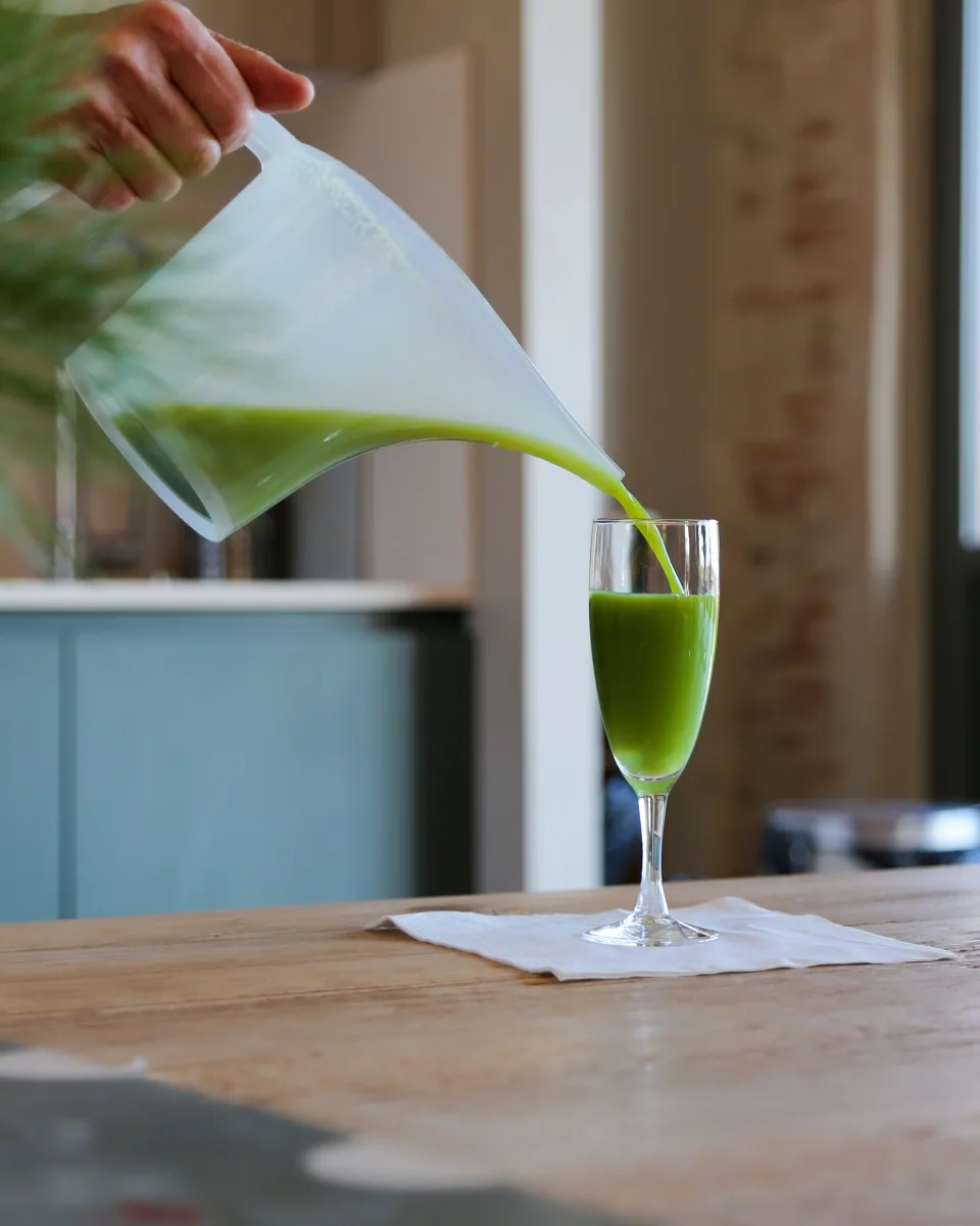 Un jus de légumes vert versé d'un pichet dans un verre, sur une table de bois