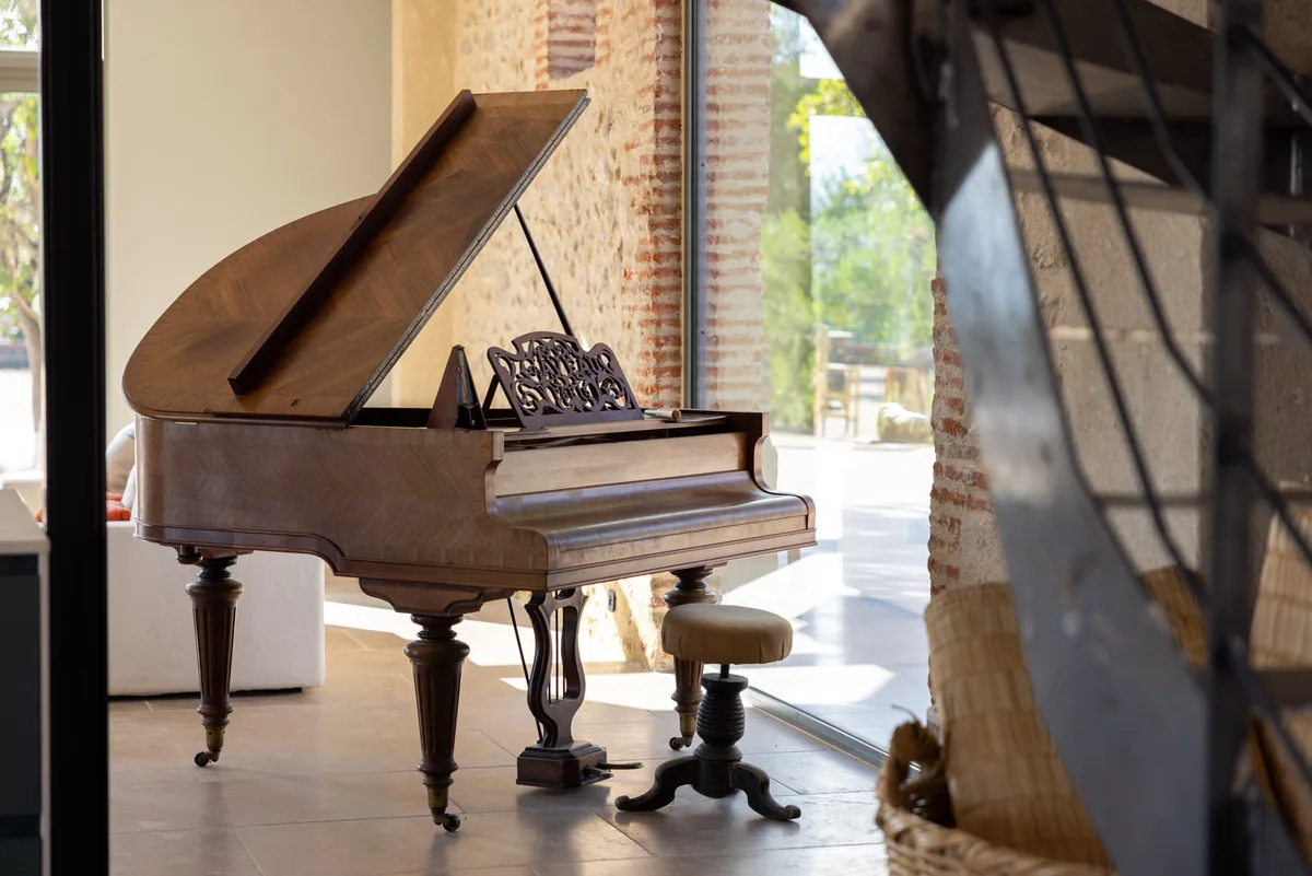 Un piano à queue et son tabouret devant une grande baie vitrée, murs de brique et de pierre