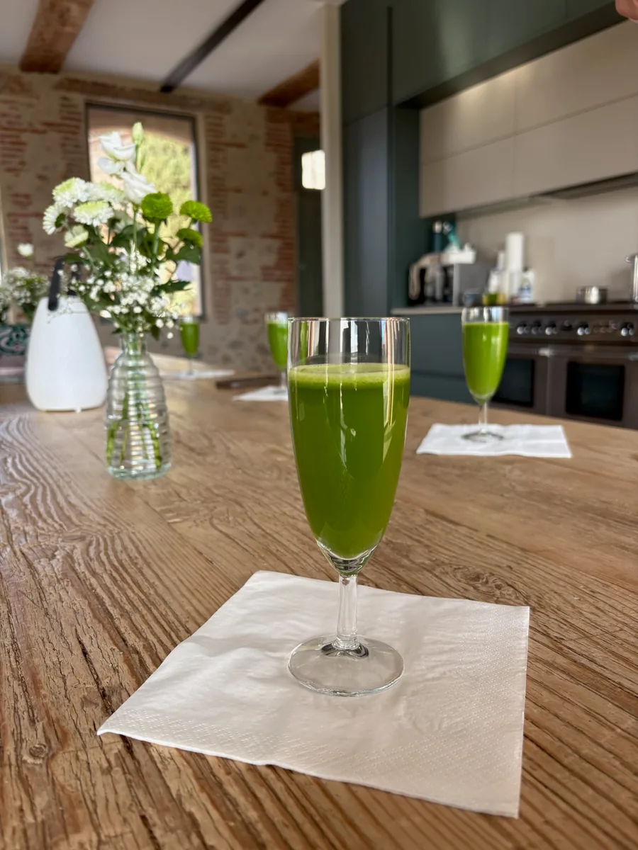 Verres à pied de jus vert alignés sur une longue table de bois, un bouquet blanc et la cuisine au fond