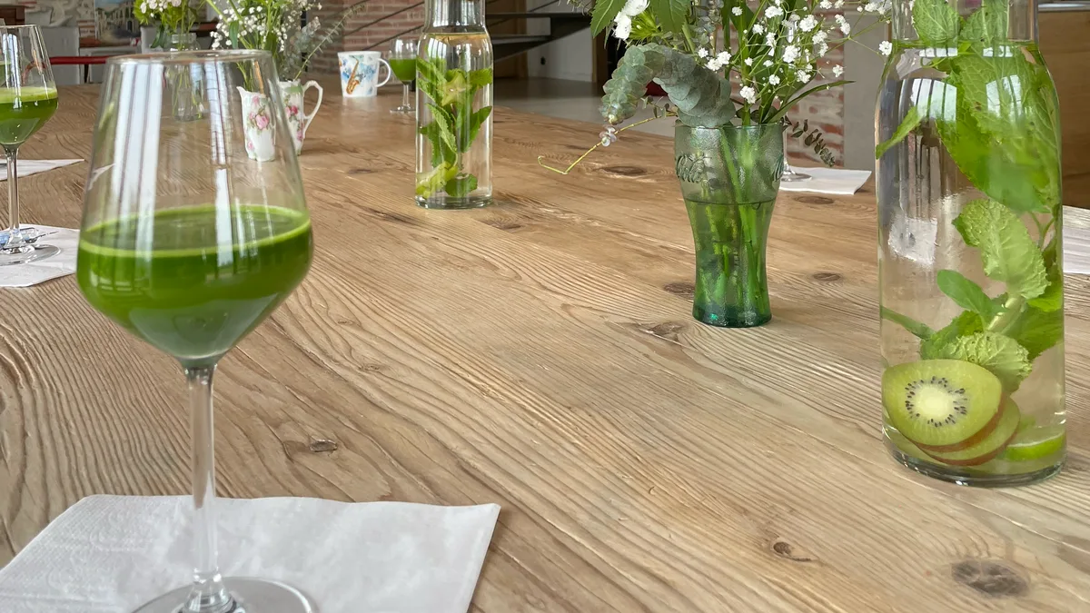 Un verre de jus de légumes vert sur une grande table en bois, à côté de carafes d'eau infusée au kiwi et à la menthe.