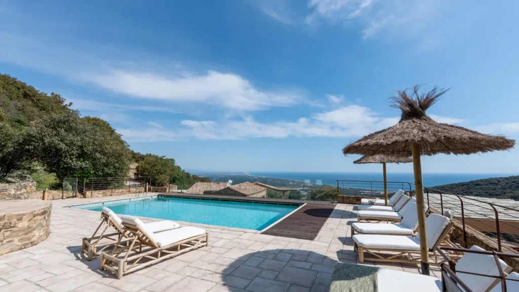 Une piscine bordée de transats et d'un parasol de paille, ouverte sur les collines et la mer à l'horizon.