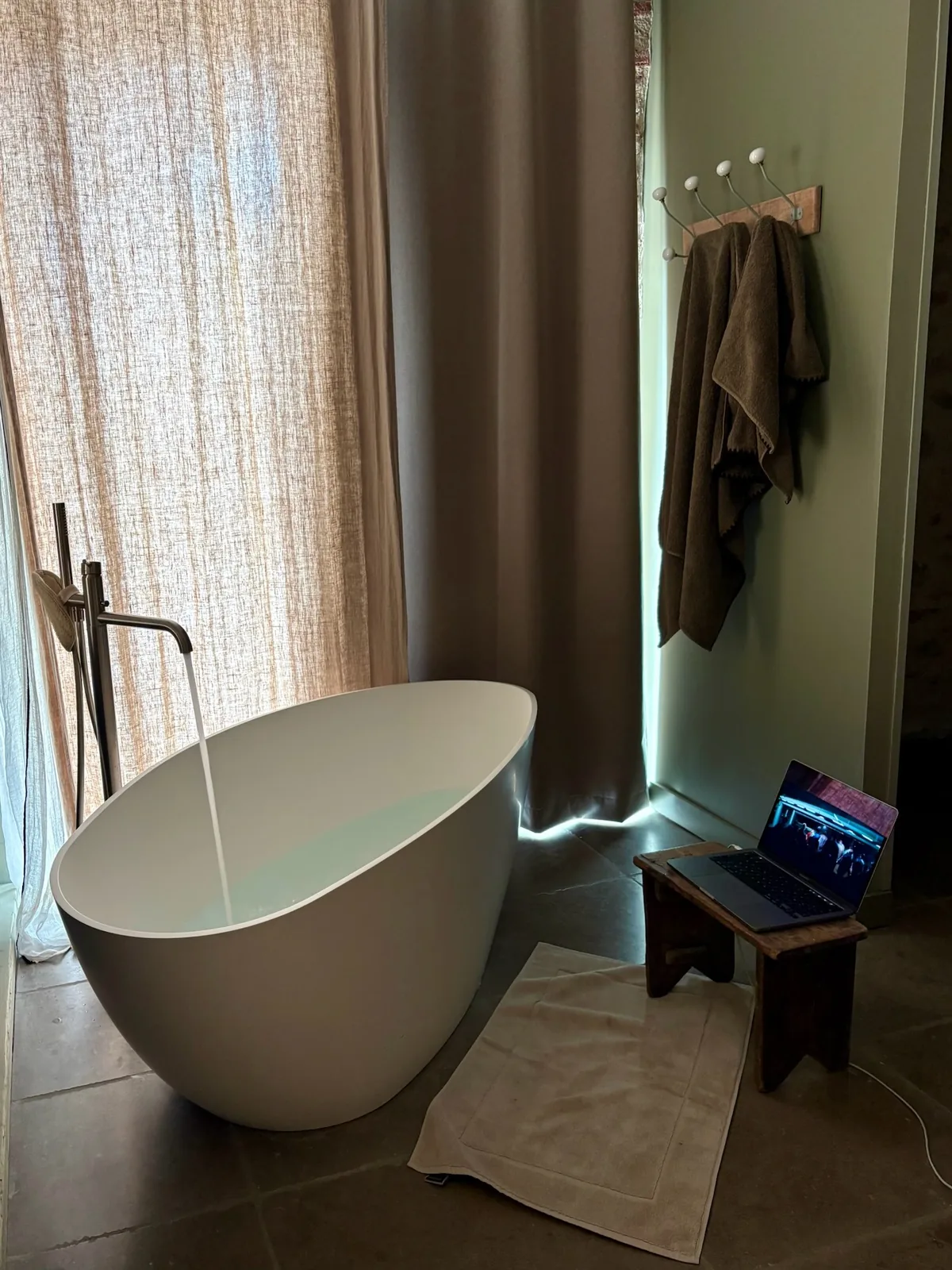 Baignoire indépendante blanche sous un robinet colonne, devant des rideaux de lin clair, dans une chambre à la lumière douce